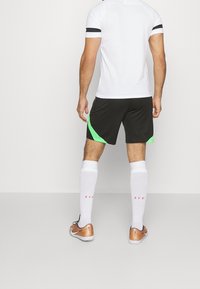 Biała koszulka sportowa z czarnymi akcentami, czarne szorty z zielonymi bokami, białe skarpetki do kolan i pomarańczowe buty piłkarskie, widok z tyłu.