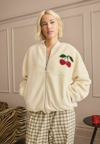 Cream fleece jacket with a zip closure and cherry embroidery. Paired with checkered beige and black trousers. Soft, textured fabric.