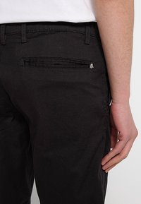Vue de profil d'une personne portant un pantalon noir avec une poche passepoilée à l'arrière et une chemise blanche rentrée, la main droite reposant le long du corps.