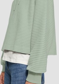 Mintgrüner, übergroßer Pullover mit gerippter Textur, Kapuze mit Kordelzug und elastischen Bündchen, über einem weißen T-Shirt und blauen Jeans getragen.