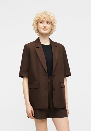 Jeune personne aux cheveux blonds bouclés portant un blazer marron à manches courtes et un short assorti par-dessus un haut noir, debout devant un fond blanc.