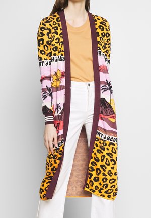 Femme portant un long cardigan à imprimé léopard, motif coucher de soleil tropical et bordure bordeaux sur une chemise moutarde et un pantalon blanc.