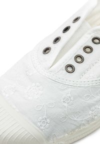 Bijela platnena tenisica s cvjetnim vezom, metalnim okovima i teksturiranom gumenom potplatom, naglašavajući jednostavan i čist dizajn.