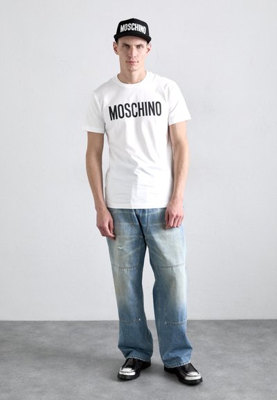 T-shirt blanche avec le logo "MOSCHINO" noir, associée à un jean large délavé bleu et des chaussures noires avec des accents argentés et une casquette assortie.