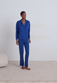 Mėlynas pižamos komplektas, kuriame yra apykaklė, su sagomis susegama marškinėliai su kišene krūtinėje ir atitinkantys siauri kelnių modeliai. Lygus audinys ir laisvas sijonas.