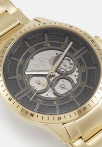 Reloj de acero inoxidable con tono dorado y una esfera texturizada negra, subesferas esqueléticas y marcadores luminiscentes. Presenta un fondo con un patrón hexagonal.