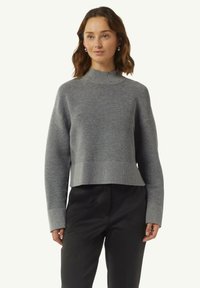 Grauer, taillierter Pullover mit einem Stehkragen, versehen mit horizontalen Streifen, Rippbündchen und einer strukturierten Oberfläche. Getragen mit schwarzen Hosen.