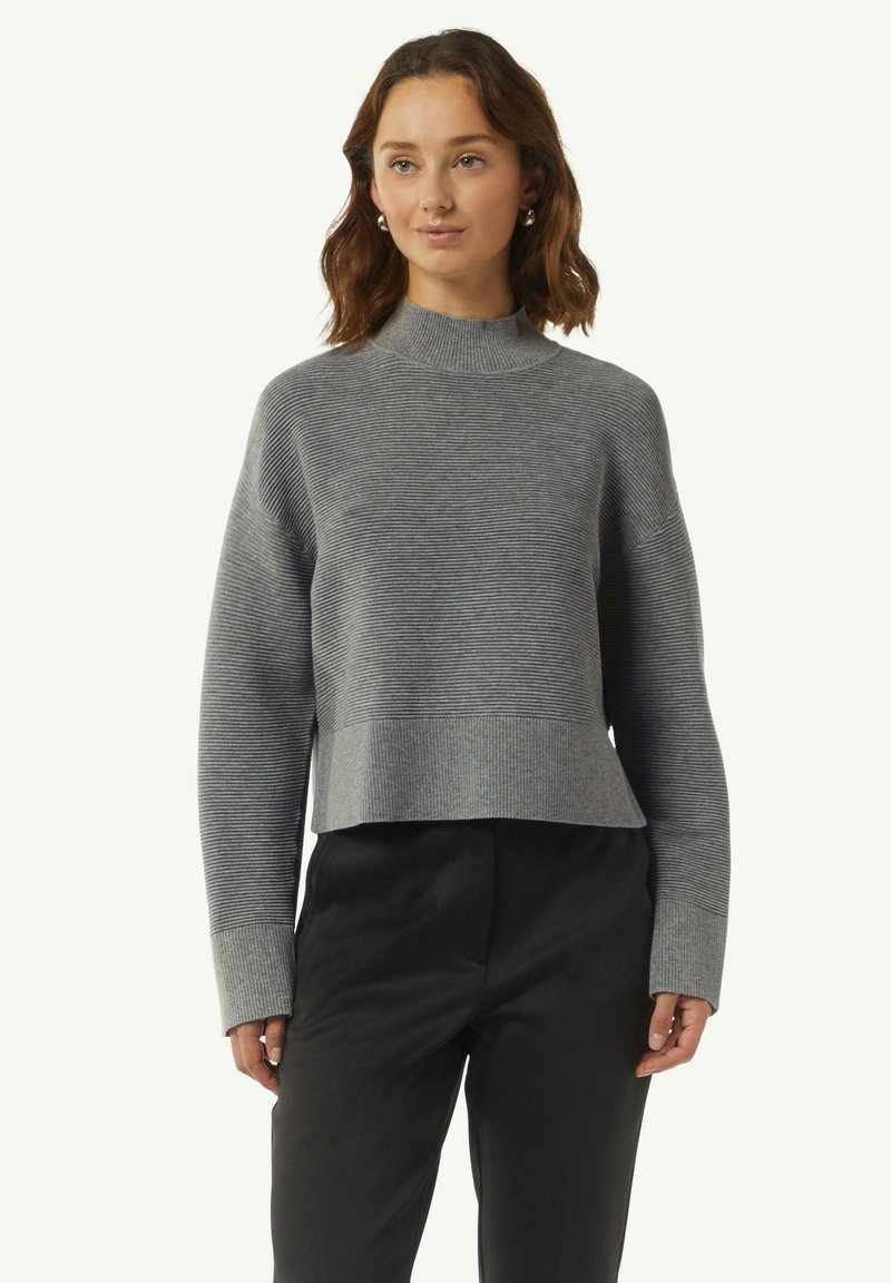 Grauer, taillierter Pullover mit einem Stehkragen, versehen mit horizontalen Streifen, Rippbündchen und einer strukturierten Oberfläche. Getragen mit schwarzen Hosen.