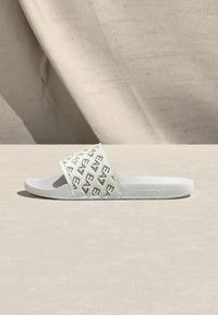 Λευκό σανδάλι slide με σόλα από καουτσούκ, διαθέτοντας λουρί με σχέδιο και μαύρες λογότυπες λεπτομέρειες, καθώς και επίπεδη, ανοιχτή σχεδίαση για εύκολη χρήση.