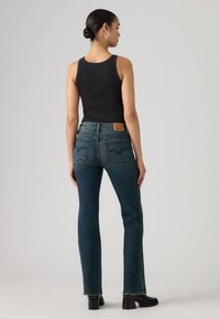 Haut noir sans manches associé à un jean droit bleu foncé. Le jean comporte des poches arrière avec des détails de couture et un patch en cuir de la marque.