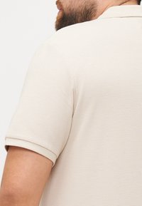 Beige polo shirt with a textured pattern, featuring short sleeves and a classic collar, showing the back view of the garment.