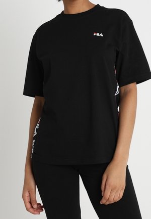 Osoba ubrana w czarną koszulkę z krótkim rękawem FILA z logiem na piersi i boku, w połączeniu z czarnymi spodniami na jednolitym tle.
