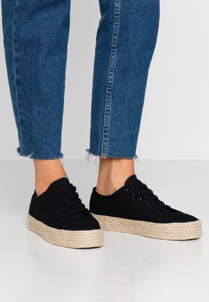 Personne portant un jean court bleu et des baskets noires à lacets avec des semelles tressées épaisses beige sur un sol clair.