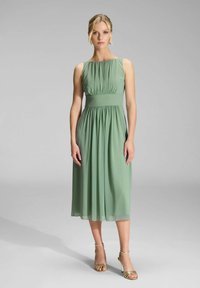 Robe en chiffon verte avec un buste plissé, longueur mi-mollet et une taille cintrée. Le mannequin porte des talons dorés à lanières.