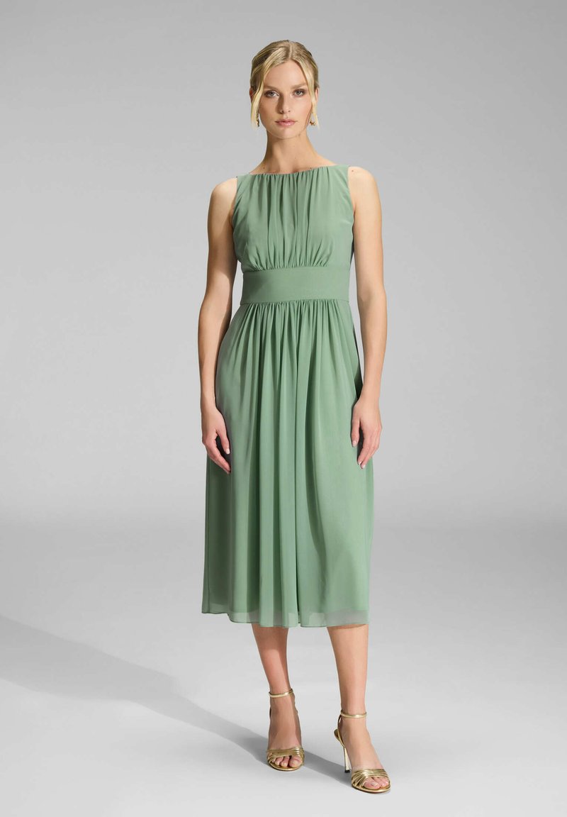 Robe en chiffon verte avec un buste plissé, longueur mi-mollet et une taille cintrée. Le mannequin porte des talons dorés à lanières.