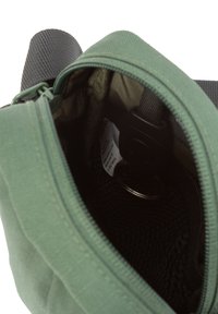 Grüne Tasche, die teilweise geöffnet ist, sodass ein schwarzes Innenleben mit einem Schlüsselring und einem sichtbaren Stoffetikett im Inneren zu erkennen ist.