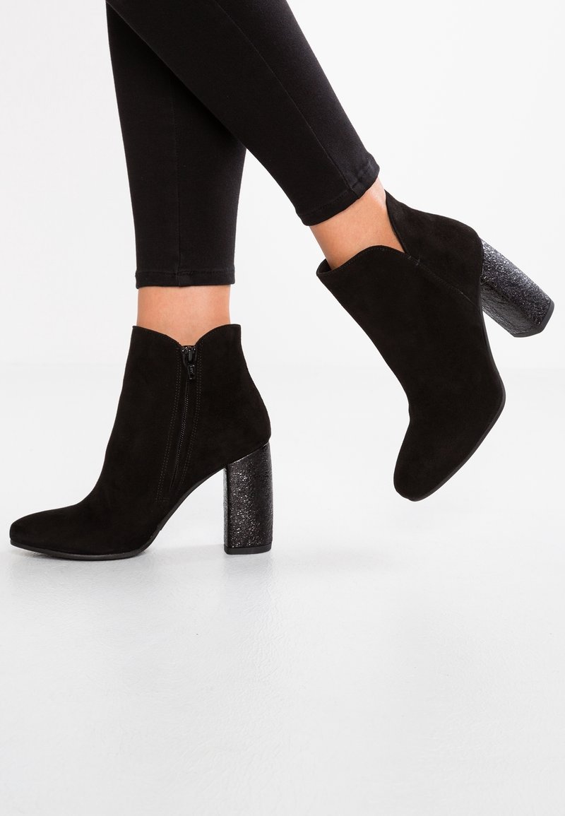 Czarne zamszowe botki za kostkę z gruboziarnistym, błyszczącym obcasem. Mają skośne cięcie oraz boczny zamek błyskawiczny dla łatwego zakładania. Gładka tekstura całości.