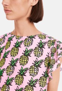 Rosa Oberteil mit einem Ananasdruck in Gelb und Grün, umrandet mit schwarzer Kontur. Rüschensleeves verleihen dem Design Textur.