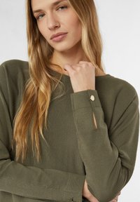 Sweter w kolorze oliwkowej zieleni wykonany z miękkiego materiału; posiada okrągły dekolt, długie rękawy z wycięciami oraz okrągłe złote akcenty na mankietach.