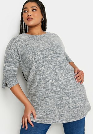 Túnica gris de manga larga con un tejido de punto texturizado, que presenta detalles de botones en los lados y un corte relajado, combinada con unos vaqueros azules.