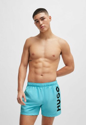 Shorts de bain en polyester bleu avec une taille élastique et un grand logo noir "HUGO" sur le côté, présentant une texture lisse et brillante.