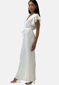 Jumpsuit bianco plissettato con maniche a volant, scollatura a V e vita con laccio. Presenta una consistenza liscia e una silhouette a gamba larga. Sandali argentati.