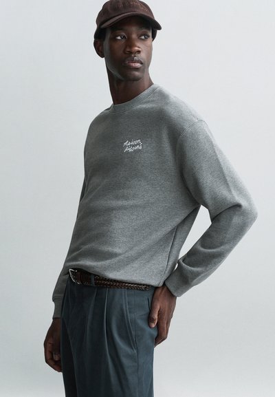 Sweatshirt gris avec un col rond et des manches longues, ornée de texte brodé, associée à un pantalon bleu foncé et une casquette marron.