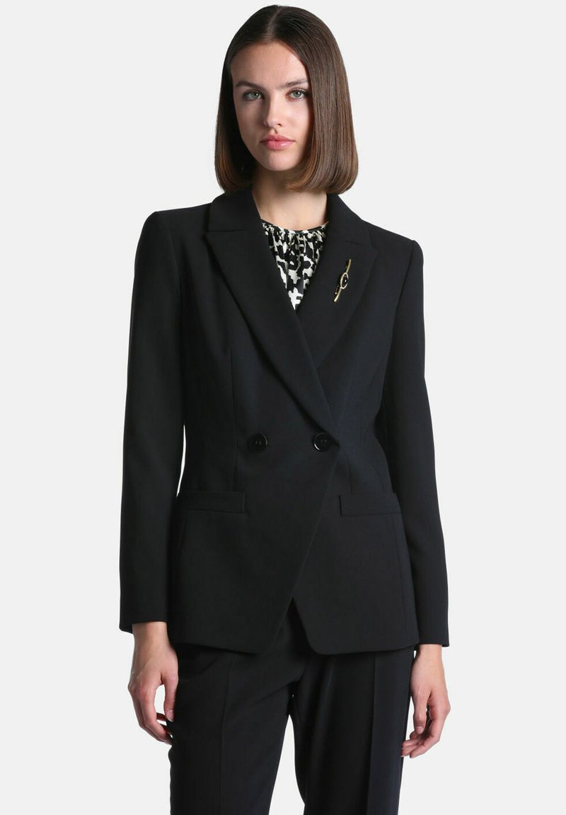 Femme aux cheveux bruns lisses portant un blazer noir croisé sur un haut à motifs et un pantalon noir assorti sur un fond uni.