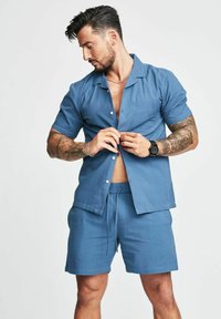 Camicetta blu a maniche corte e pantaloni coordinati realizzati in tessuto testurizzato. La camicia presenta una chiusura a bottoni e un design sobrio e rilassato.