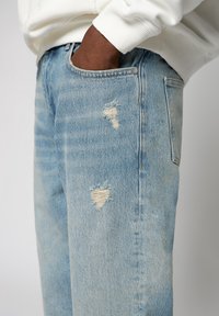 Jean en denim bleu clair avec des pièces usées aux bords effilochés. Le tissu présente un aspect délavé et texturé, témoignant d'une usure apparente.