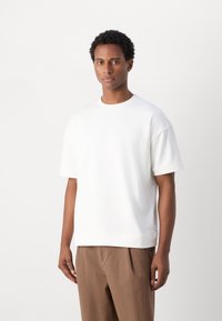 Bílý oversized tričko vyrobené z měkké, hladké látky s krátkými rukávy a kulatým výstřihem, doplněné o volné hnědé kalhoty.