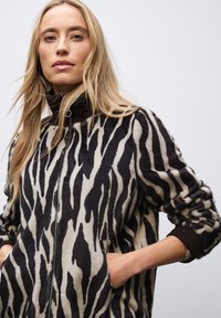 Veste imprimée zèbre avec fermeture éclair à l'avant, fabriquée en tissu doux. Dotée d'un col montant et de poches latérales ; les couleurs incluent le noir et la crème.