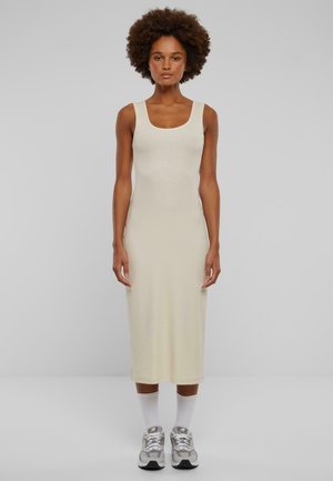 Beige ribbed knit tank dress, mid-length, featuring a square neckline and a fitted silhouette. Paired with white socks and grey sneakers.