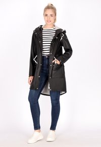 Cappotto impermeabile nero con cordini, accessori in argento e tasche frontali, abbinato a una maglietta a righe e jeans blu. Sneakers bianche.