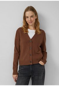 Brązowy sweter kardigan z dzianiny z dekoltem w serek, ściągaczami na rękawach i u dołu oraz pięcioma guzikami z przodu. Nosi się go na białą koszulkę.