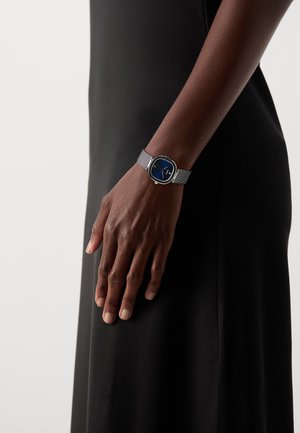 Montre ronde avec un cadran bleu, des éléments en argent et un bracelet en maille texturée, portée sur une main contre un vêtement noir.