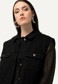Schwarze Jeansweste mit großen Brusttaschen, silbernen Metallteilen und kragenlosem Ausschnitt, über einem schwarz gestreiften Mesh-Top getragen.