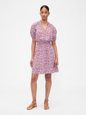 Woman standing wearing a short-sleeve, pink floral dress with a smocked waist and brown strappy flat sandals on a white background.
