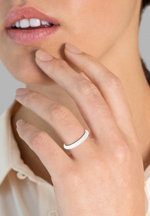 Anello bianco e liscio indossato su un dito, caratterizzato da un design minimalista. La mano tocca delicatamente il mento su uno sfondo neutro.
