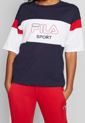 Osoba ubrana w granatowo-biało-czerwoną koszulkę FILA Sport oraz czerwone spodnie, z rękami w kieszeniach, stojąca na tle jasnoszarego tła.