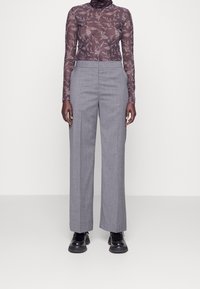 Långärmad, figurnära genomskinlig topp i mörk lila med blommönster, kombinerad med höga, ljusgrå vida byxor och svarta skor.