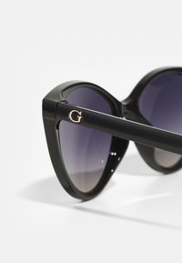 Lunettes de soleil noires avec une forme œil de chat, verres dégradés violets et un accent doré en forme de 'G' sur la branche. Texture de monture en plastique lisse.