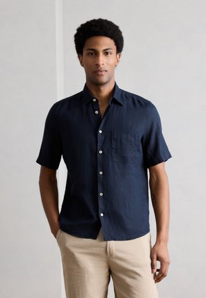 Άνδρας που στέκεται με το σώμα του προς τα εμπρός, φοράει μπλε κοντομάνικο πουκάμισο με κουμπιά και μπεζ παντελόνι, με τα χέρια μερικώς στις τσέπες, ουδέτερο φόντο.