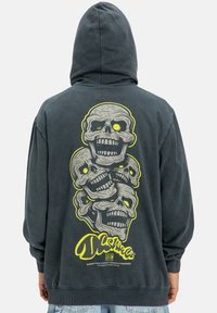 Tmavohnedá mikina s kapucňou. Na zadnej strane je grafika štyroch lebiek v žltej a sivej farbe. Čierne a žlté logo značky na spodnej strane. Materiál: bavlna.