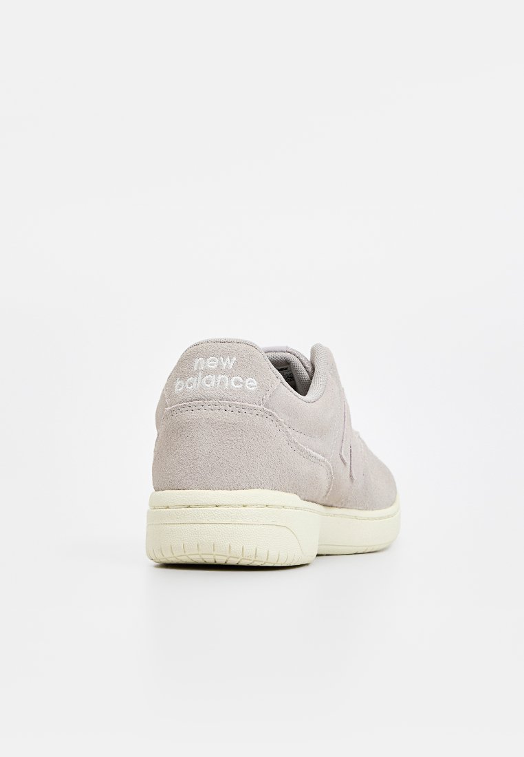 Zapatilla de gamuza gris clara con diseño de corte bajo, que presenta una superficie texturizada, suela de goma color crema y logo de "new balance" en el talón.