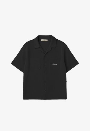 Junge Frau trägt ein schwarzes Hemd mit Knöpfen und dem Schriftzug „Prohibited“ sowie eine schwarze Kappe mit orangem „NY“-Logo, steht vor einem grauen Hintergrund.