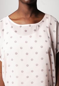 Blusa transparente en rosa pálido con cuello redondo, que presenta lunares a rayas horizontales en gris, mangas cortas y un corte suelto.