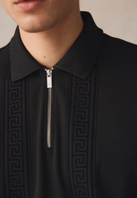 Černá zipová polo s texturovaným límcem, s vzorem na přední části a stříbrným zipovým detailem na krku.
