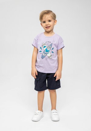 Un t-shirt court à manches courtes de couleur violet clair avec un graphique représentant des planches de surf et du texte. Associé à un short cargo bleu marine et des baskets blanches.