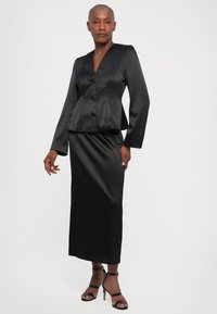 Zwarte satijnen blazer met V-hals, knopen aan de voorkant en lange mouwen, gecombineerd met een bijpassende zwarte satijnen maxi rok en strappy heels.